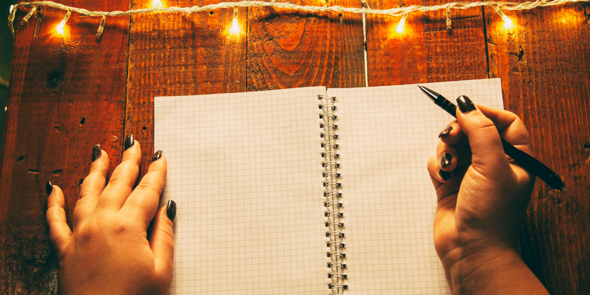 hands with a pen hovered over a graphing paper notebook with lit string lights around it