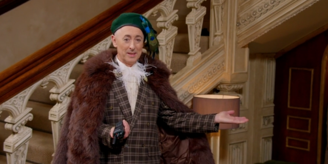 Alan Cumming, host of The Traitors Season 2, stands at a podium before an ornate staircase, raising his hands in feigned exasperation, saying, “I mean, come on.”