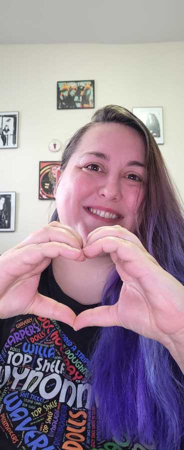 valerie anne, a white nonbinary human with purple hair, holds up her hands in a heart shape
