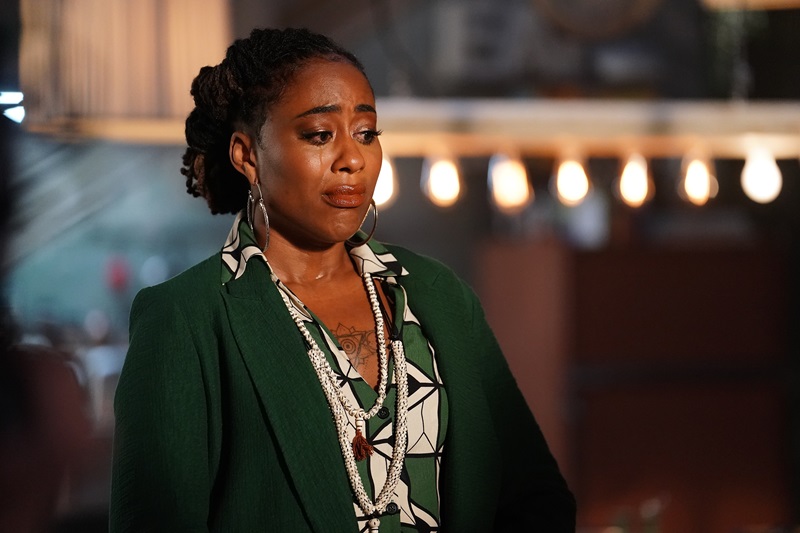Malika cries as she has to tell Isaac to leave the Coterie for his own good. She's wearing a green printed blouse and matching green blazers. Tears streak down the right side of her face.