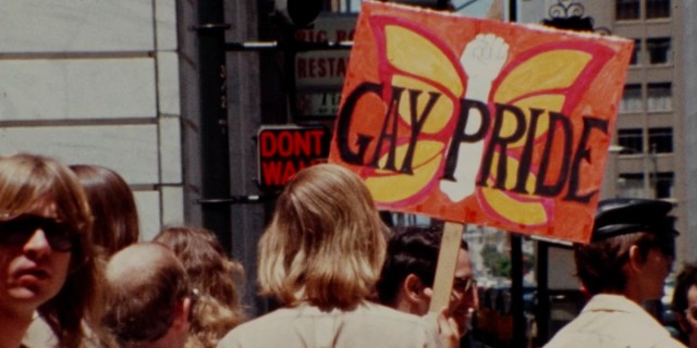 Gay USA: a still from footage from a 1977 pride event. A sign with a butterfly is held up that says Gay Pride