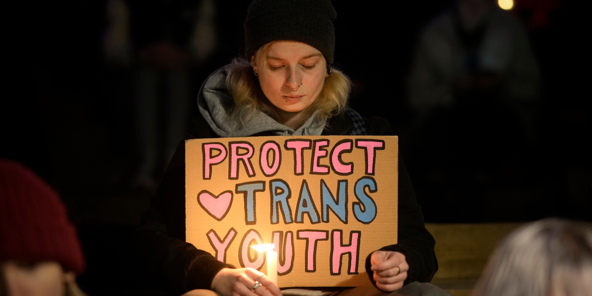 A young person at a vigil for Brianna Ghey holds a candle in one hand a sign that says Protect Trans Youth in the other
