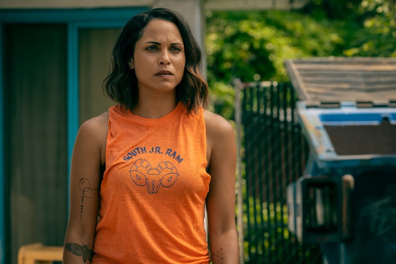 Jackie stands in a motel parking lot, in front of the dumpster she just crawled out of, watching Emma walk away. She's wearing an orange sleeveless top with South Jr. Ram on it. 