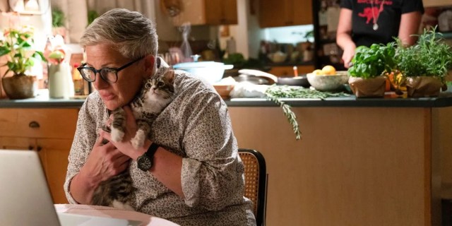 Velma (Abby McEnany) clutches a kitten while examining information on her laptop, while her wife, Teddi (Robin Weigert), cuts vegetables nearby in the kitchen.