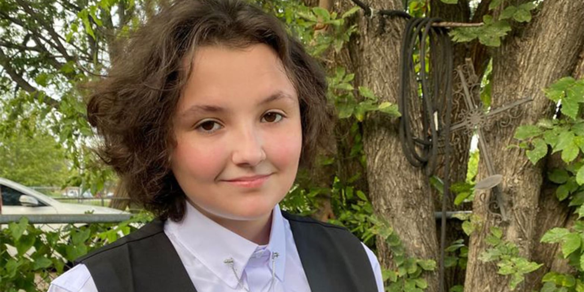 Nex Benedict, a trans teen with wavy brown hair white button down and black vest, smiles at the camera.