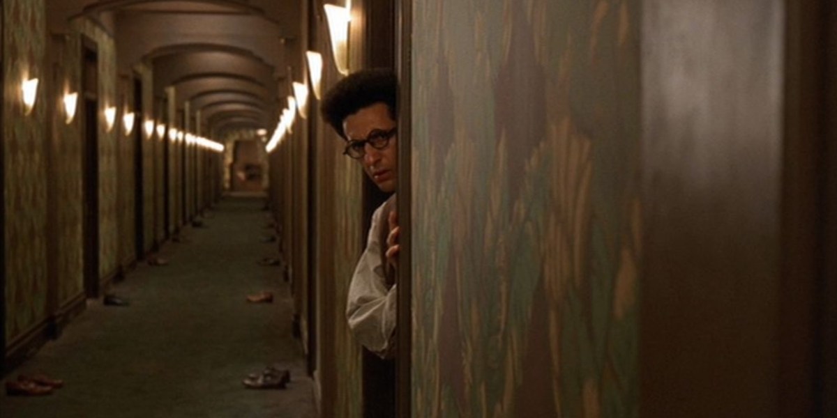 John Turturro peaks behind a door frame into a long hallway in Barton Fink