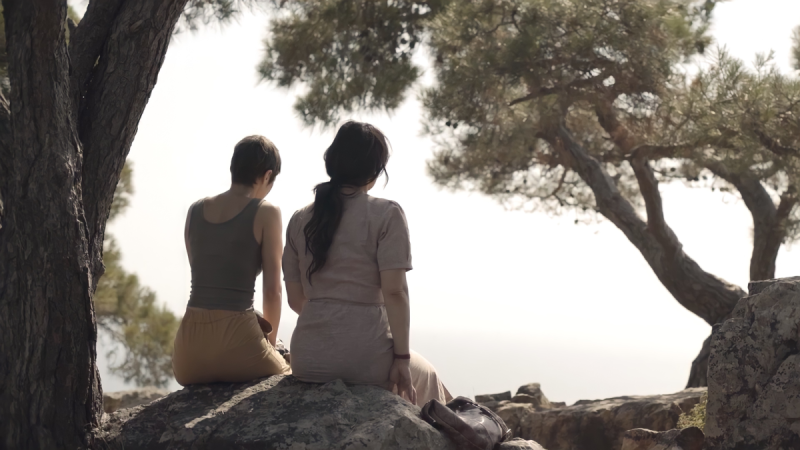 The silhouette of two women sitting on a cliff with overhanging trees and broad, white sky