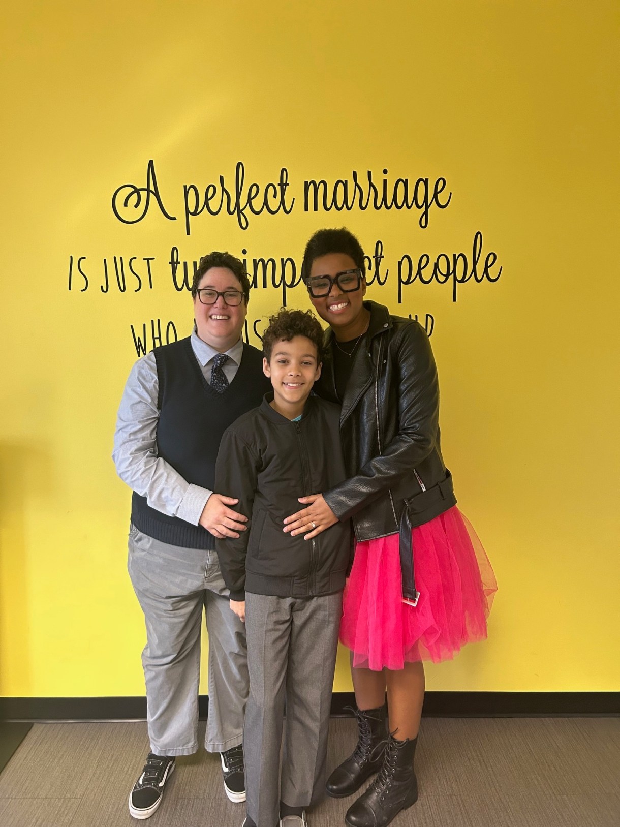 The author (a black woman with short hair) and her wife (a white butch with brown hair) pose against a yellow and green wall on their wedding day with their son.