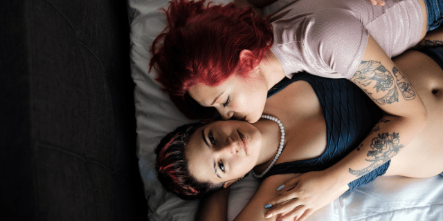 an aerial shot of two lesbians in bed, one with long dyed red hair and one with short hair with red streaks