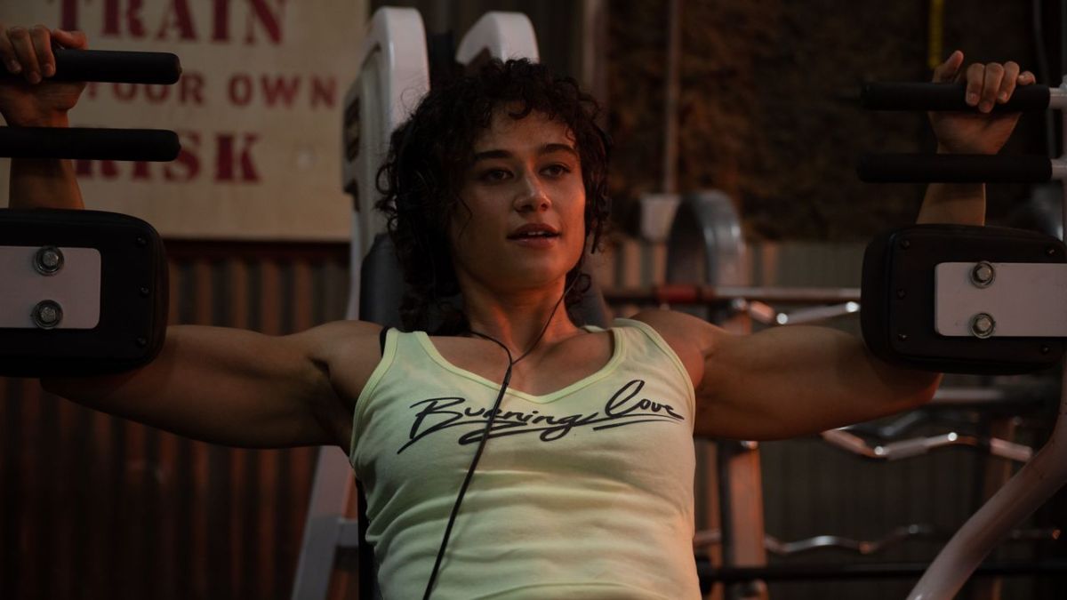 Katy O'Brian as Jacky in Love Lies Bleeding. Her muscly arms are stretched out wide on an exercise machine. She wears a white tank top.