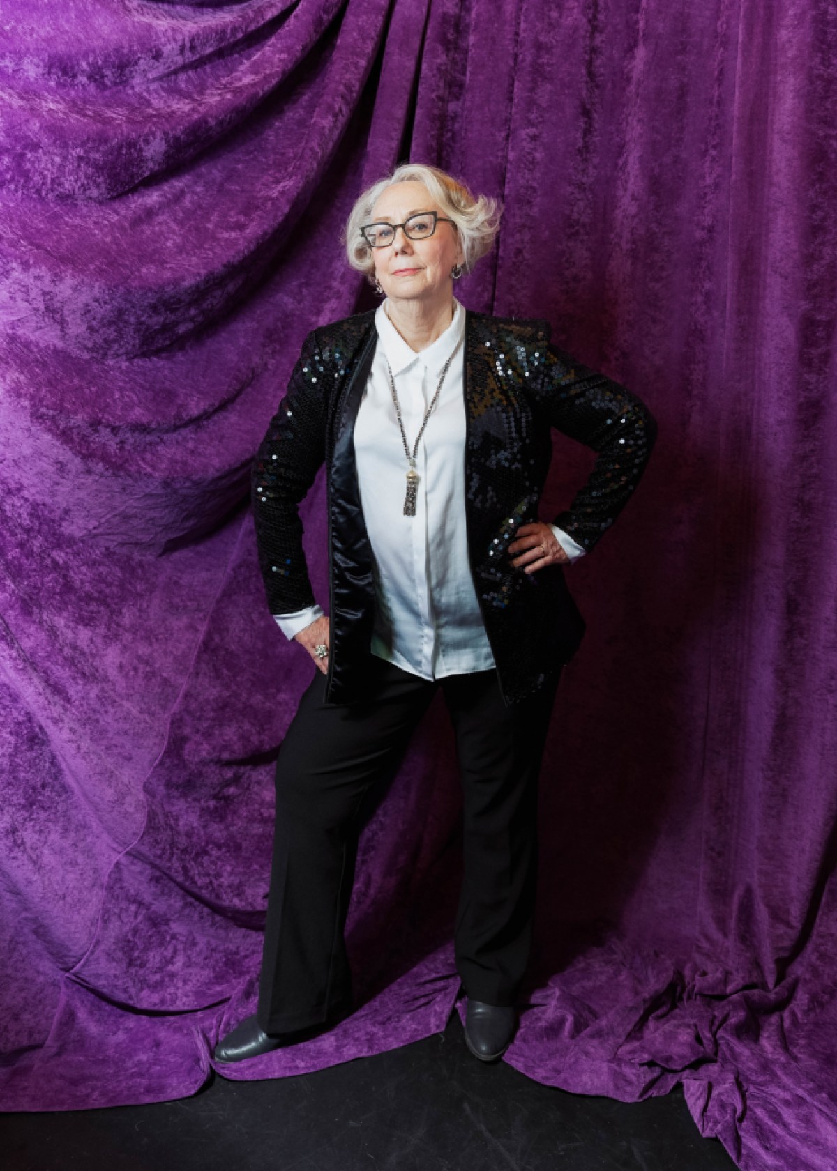 Mink Stole poses in front of a purple backdrop in a sparkly black suit