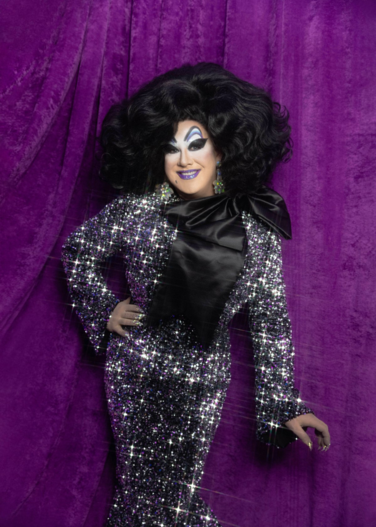 Peaches Christ in a sparkling black dress poses in front of a purple background