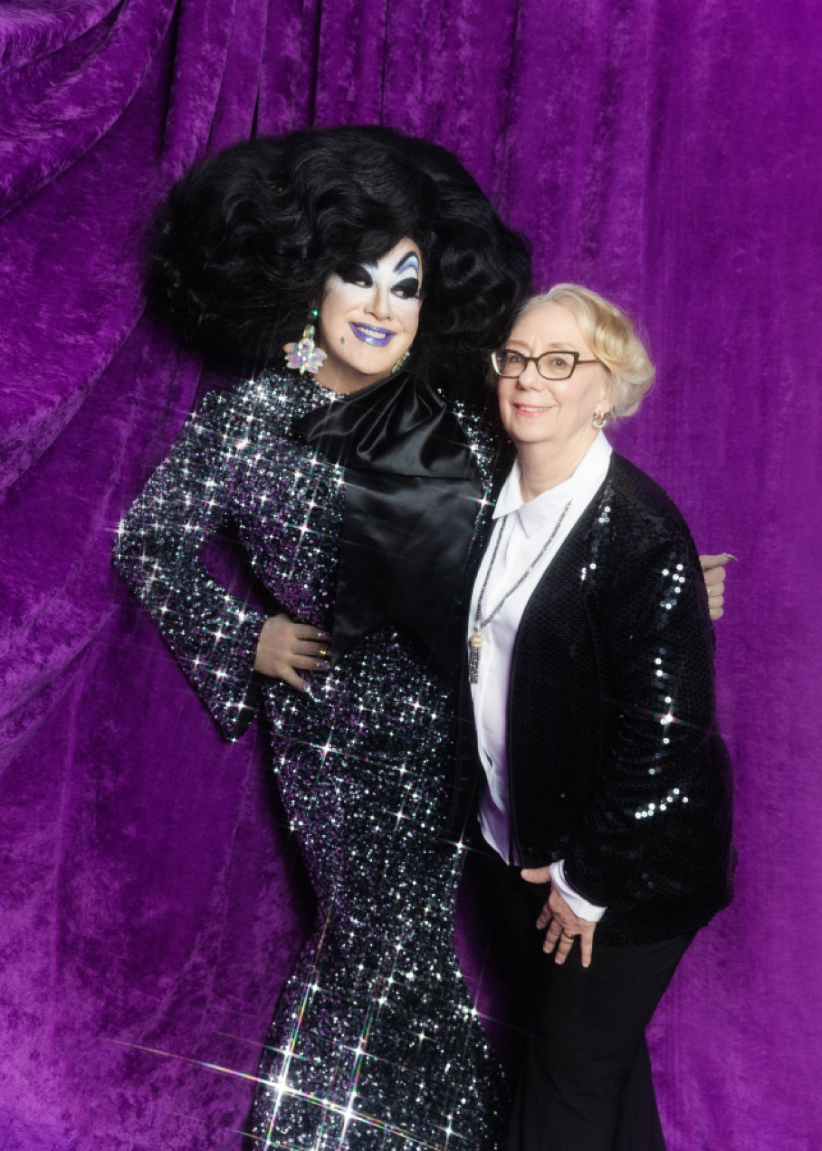 Peaches Christ Mink Stole pose in front of a purple backdrop, Peaches is in sparkly black dress, Mink is in a sparkly black suit