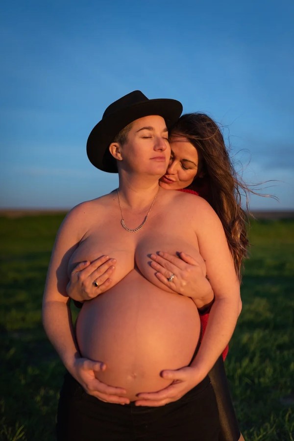 a woman stands behind a pregnant butch wearing a cowboy hat, hands covering breasts