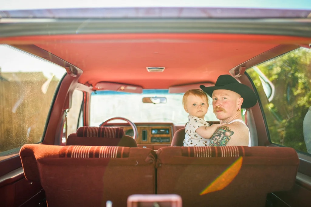a queer parent in a cowboy hat holds a baby in a retro car