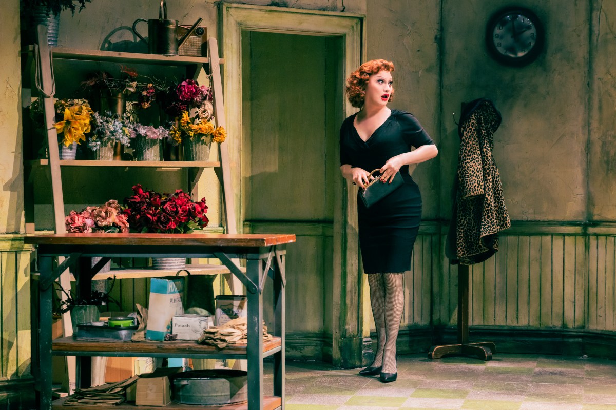 Jinkx Monsoon in Little Shop of Horrors as Audrey in a tight black dress looks over her shoulder in Mushnik's flower shop.