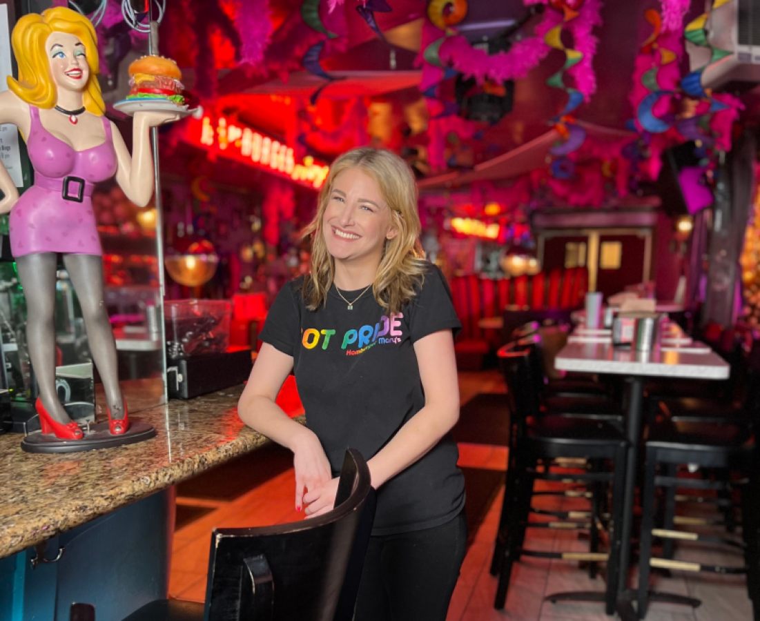 Cheyanne Dunn smiles, leaning on the bar at Hamburger Mary's next to a smiling Hamburger Mary's statue.