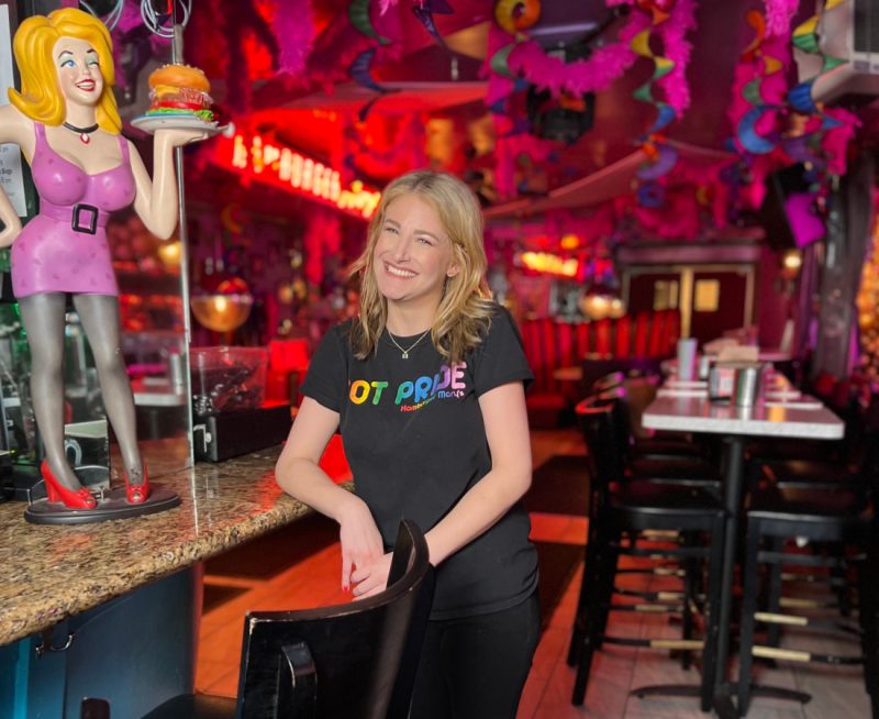 Cheyanne Dunn smiles, leaning on the bar at Hamburger Mary's next to a smiling Hamburger Mary's statue.