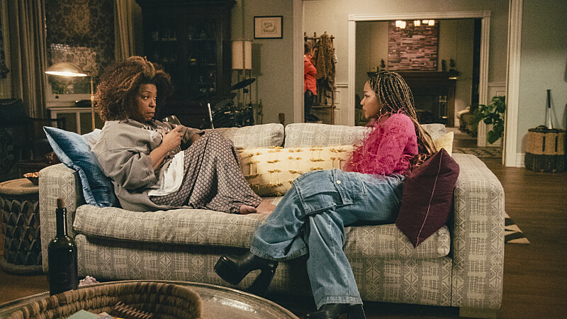 Aunt V and Delilah sit facing each other from opposite ends of the couch. Vi is in pajamas and a robe and holding a wine glass in her right hand. Vi is on the left side of the couch. Delilah is on the right side of the couch and she's wearing a feathery pink sweater, wide leg denim cargo pants and chunky black boots.