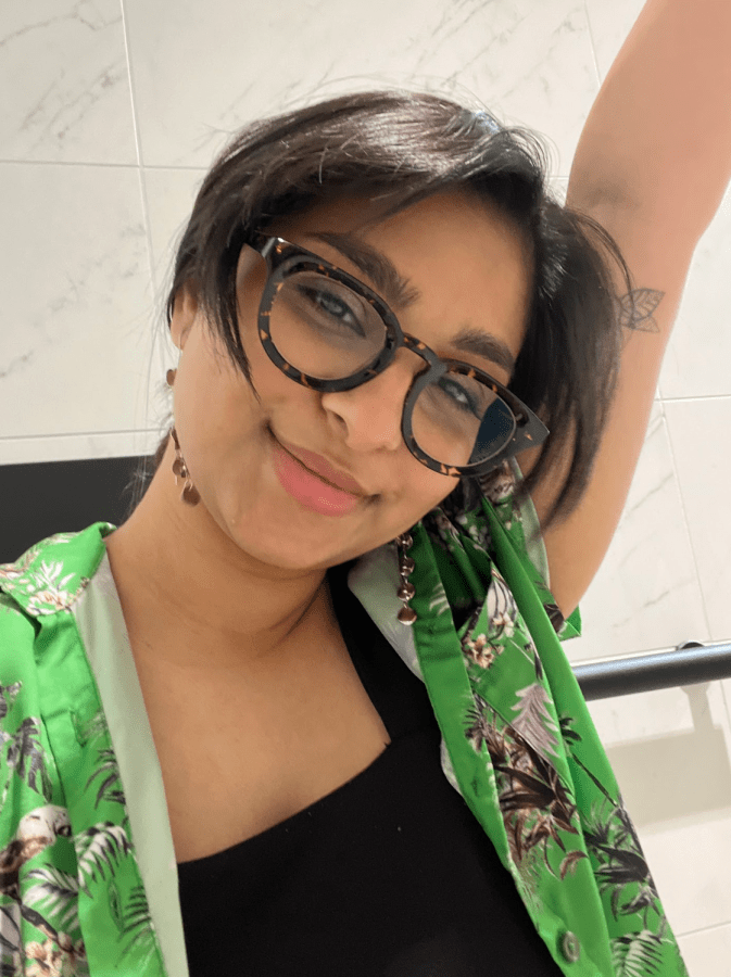 Ashni, a South Asian person, is raising an arm in this photo and smiling playfully. She has short straight dark hair and glasses and her shirt is a colorful button up.