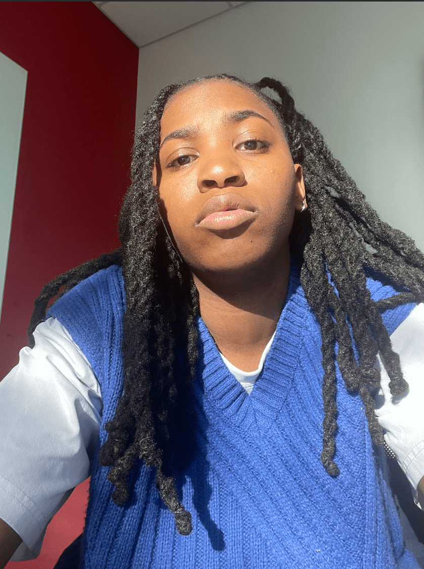 A photo of Tima a Black woman with long locs wearing a tee shirt and sweater vest looks down at the camera