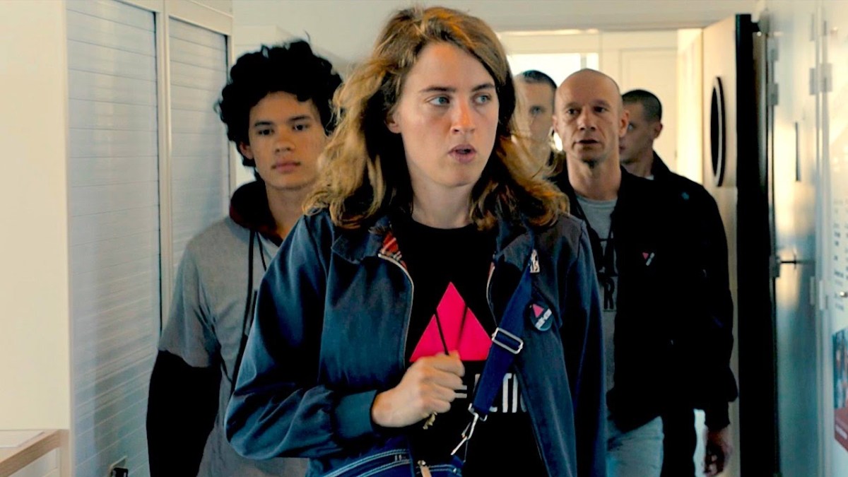 Adèle Haenel wearing an Act Up Silence=Death shirt walks through a hallway with a group of men behind her. 