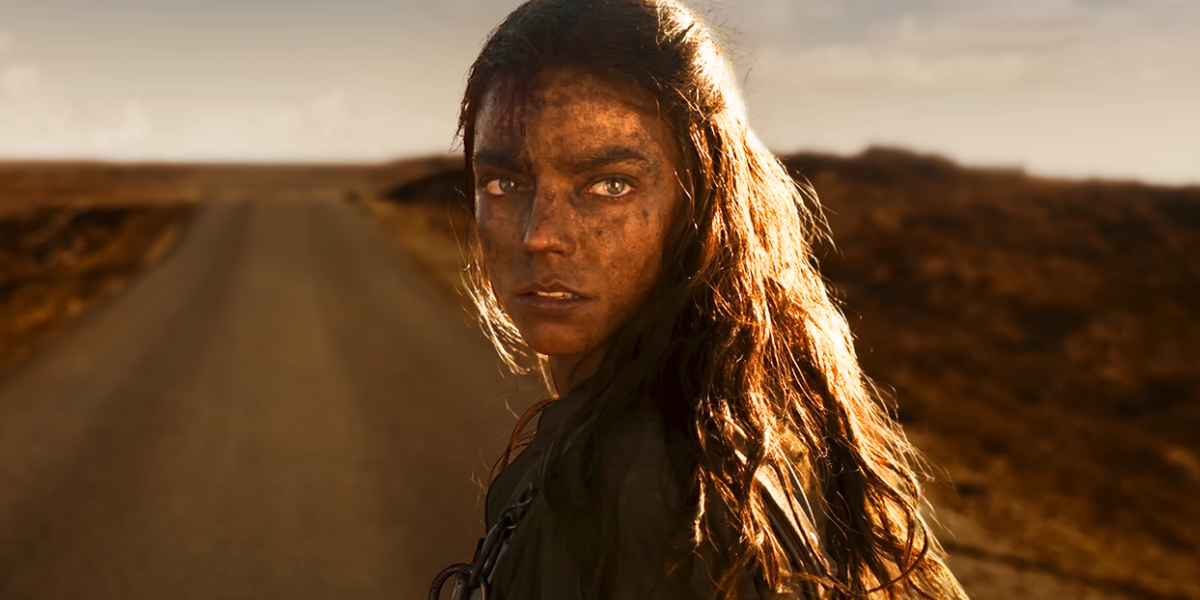 Furiosa queer: Anya Taylor Joy as Furiosa covered in dirt looks back, an open road stretching beyond her.
