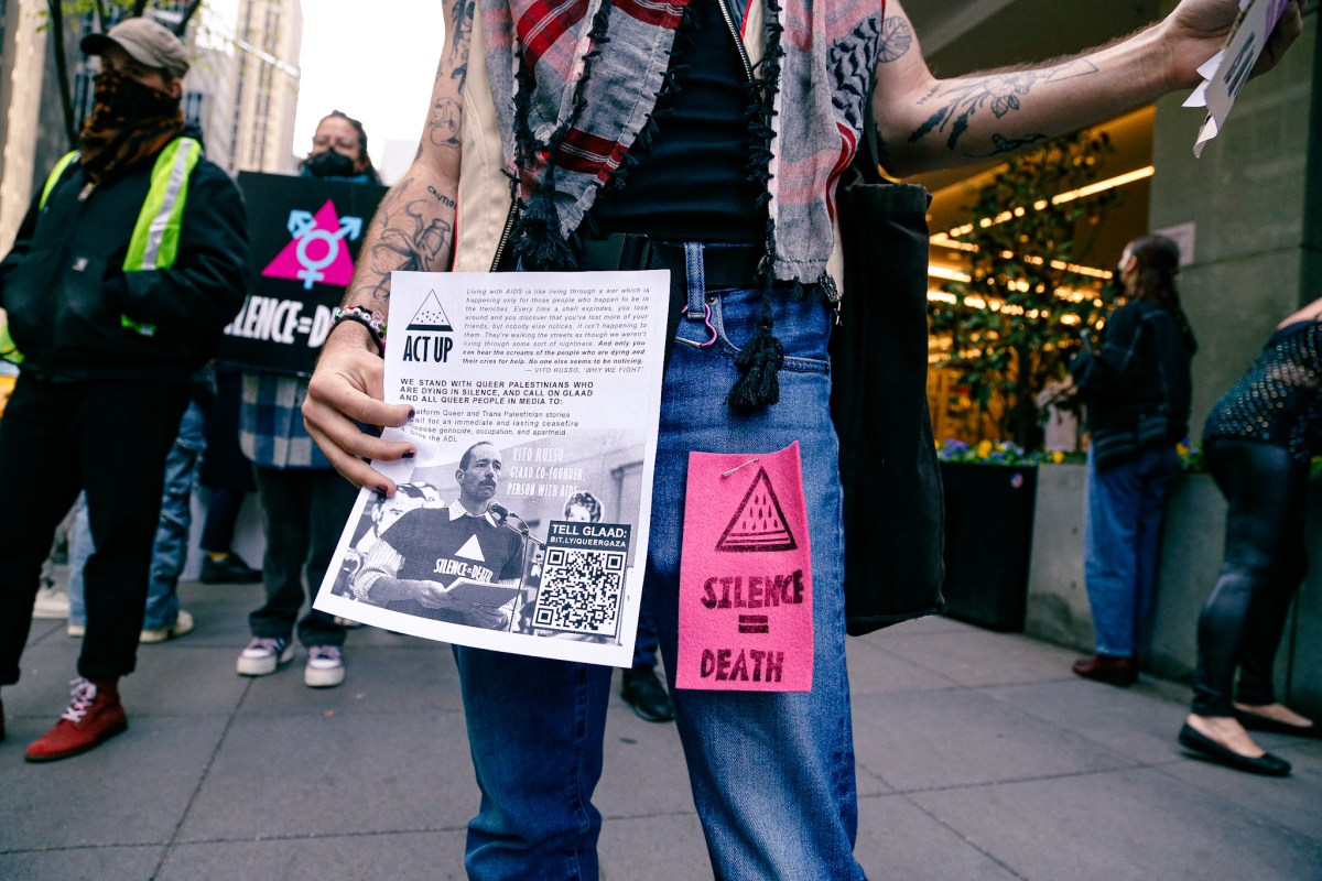 A protestor holds a flier with Vito Russo on it and wears a pinned message that says SILENCE = DEATH
