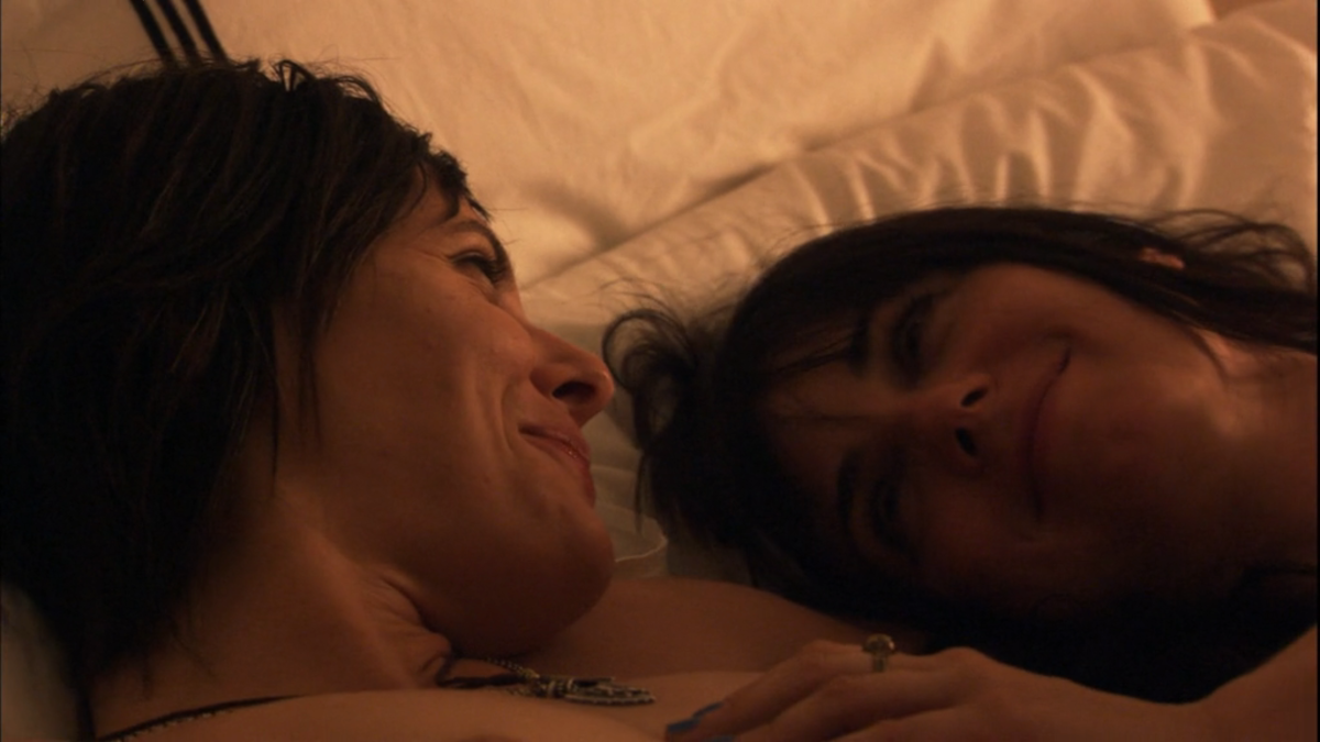 Kate Moennig and Mia Kirshner smile at each other in bed.