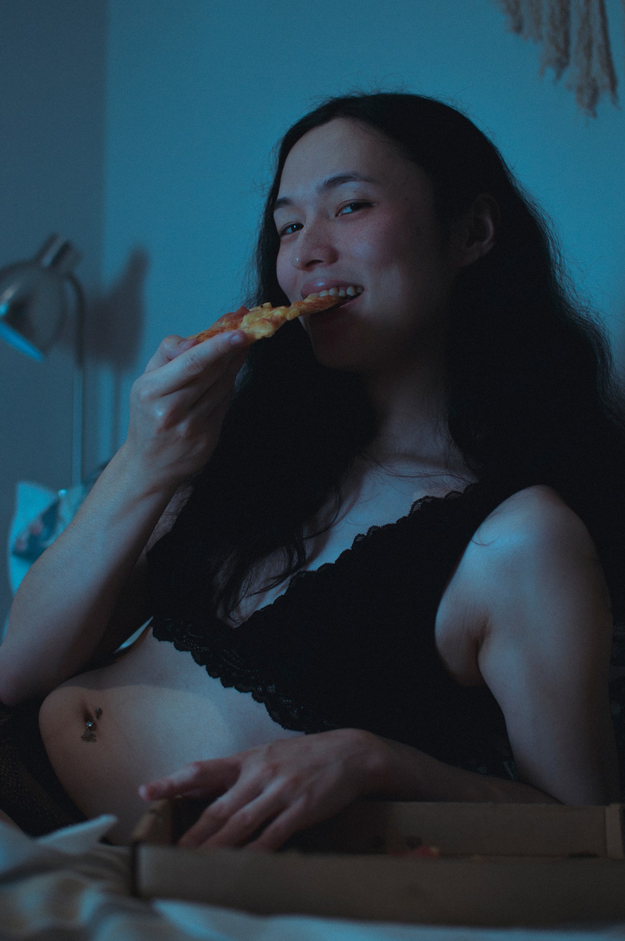 a person eats pizza in bed in a black bra while smiling at camera