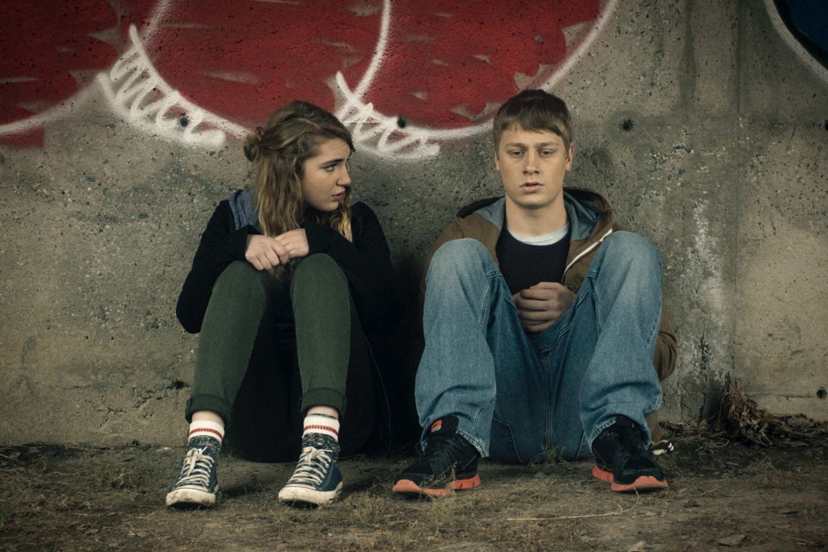 Sophie Nélisse sits against a wall next to a sad looking teen boy.