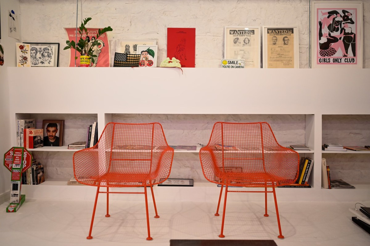 The lobby at Cake with two metal orange chairs and pro-trans signs, tattoo books, and a poster for Shakedown among other knick-knacks. 