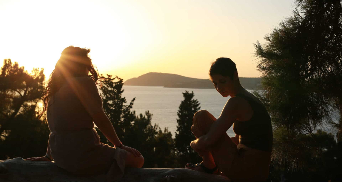 Two women sit by water silhouetted by the sunset in Love, Spells and All That