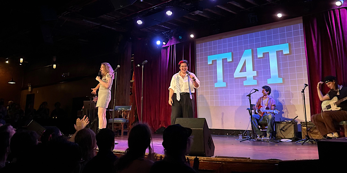T4T 3rd Anniversary Spectacular: two trans comedians stand on-stage in front of two musicians and a screen that says T4T