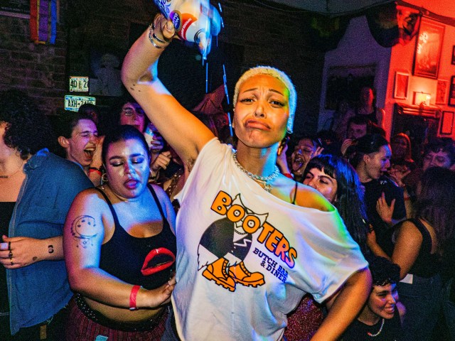 A crowd in the back room at a lesbian bar, with a person wearing a Booters white tee holding a can of whipped cream upside down.