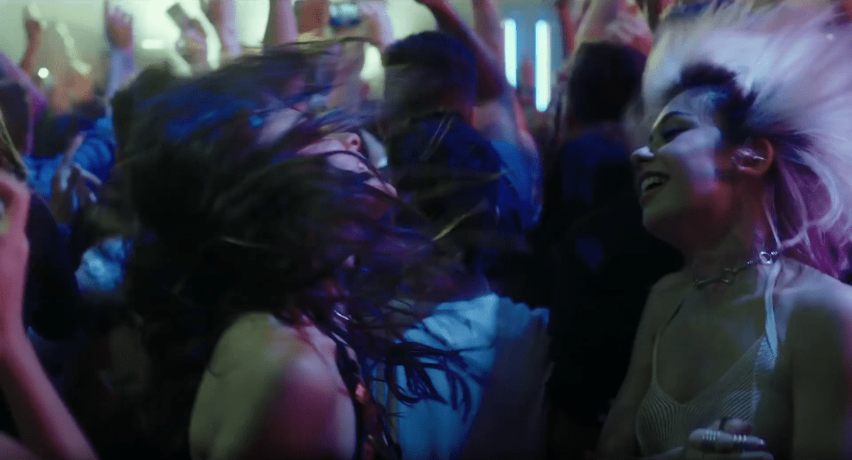 Two girls dance in a club with their hair flipped up in Holy Camp!