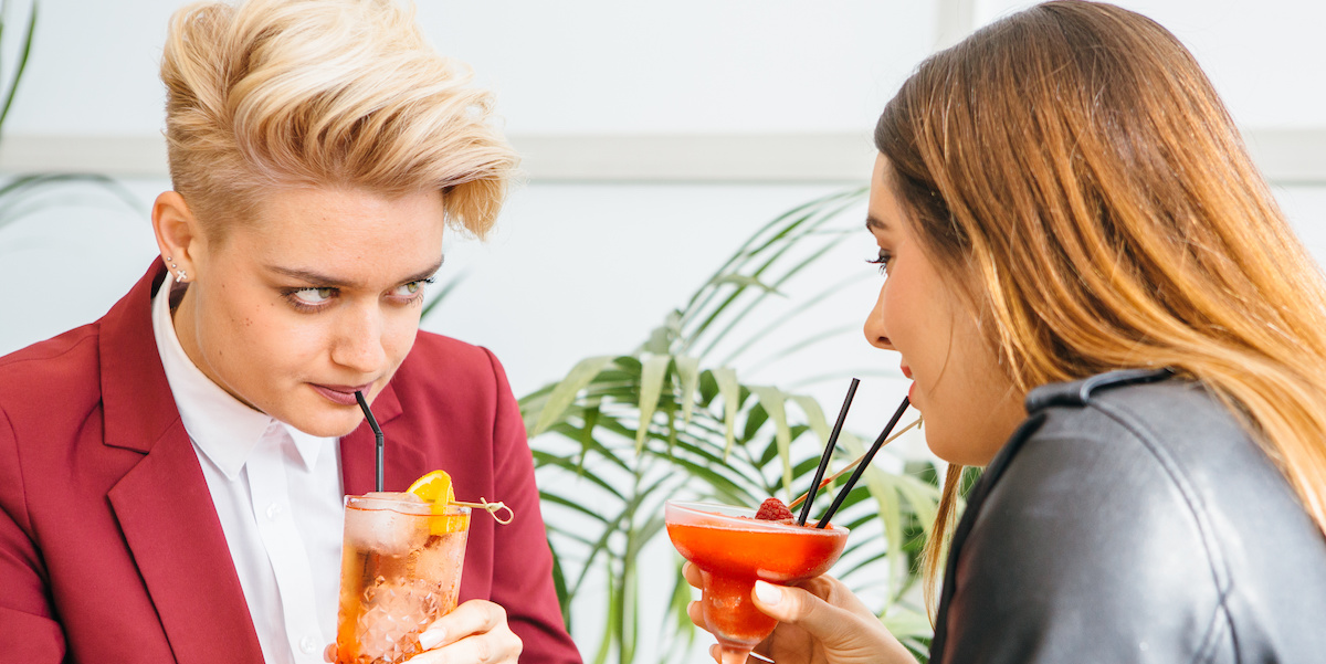 Beautiful girlfriends drinking cocktails while sitting at table in cafe and looking at each other.