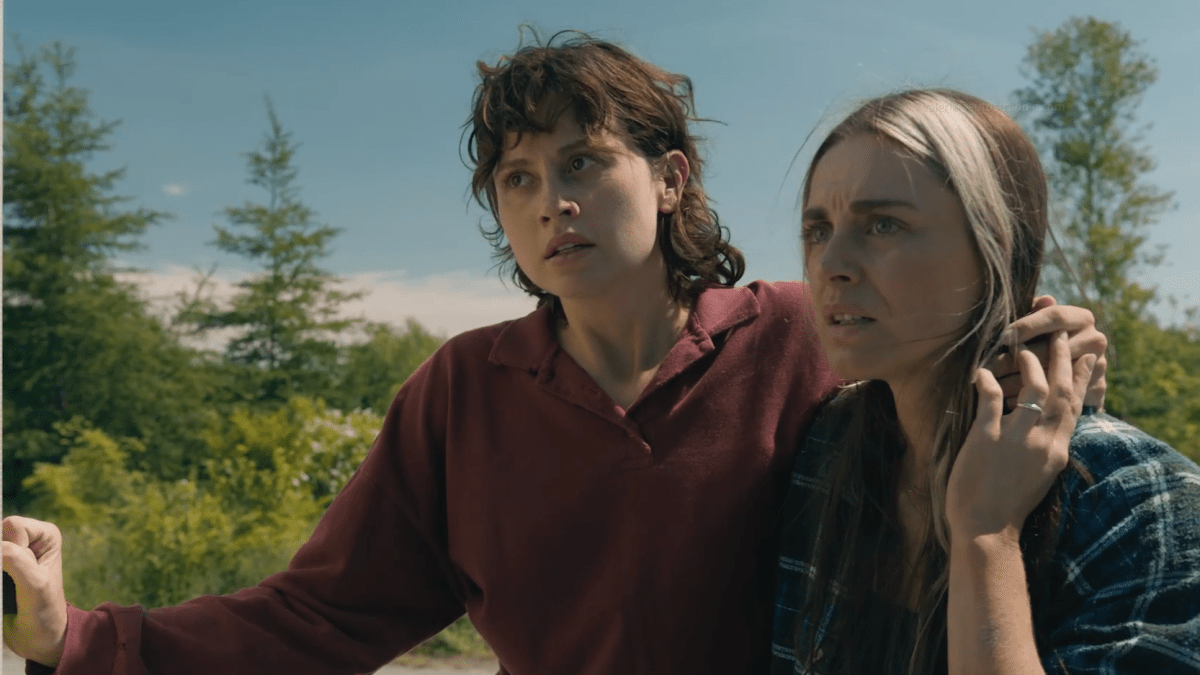 Screenshot from the TV show FROM: Kristi stands with her arm around Mari's neck, Mari holds Kristi's hand that is over her shoulder. They are both looking off screen sternly.